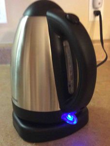 Picture of the Farberware 104556 stainless steel tea kettle, operating. Shows the blue LED pilot lamp glowing, and the water gage adjacent to the handle.