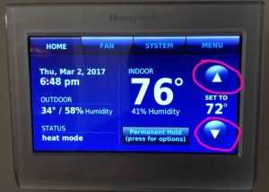 Picture of a common Honeywell thermostat, displaying its Home screen, with the temperature adjustment buttons circled.