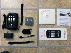 Picture of the HumanWare Victor Reader Trek navigator, showing the contents of the original box, Including the Trek, USB AC power adapter, long and short USB cables, wrist and shoulder straps, earbuds, manual, warranty paper, and DVD disc.