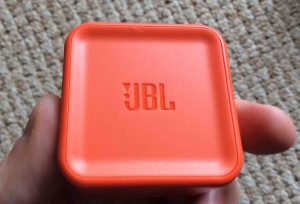 Picture of the JBL Charge 3 waterproof rechargeable speaker, showing its USB power supply adapter, front view, held in hand.