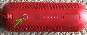 Picture of a common Bluetooth speaker, top view, power button glowing white, highlighted. Speaker powered ON. 