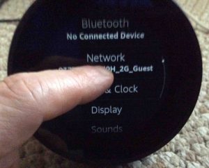 Picture of the Amazon Echo Spot speaker, displaying its Settings screen. Showing a finger swiping up to scroll down through the list of settings.