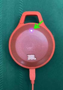 Picture of the JBL Clip speaker, powered ON while charging, showing the purple battery indicator lamp.