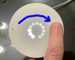 Picture of the top of the Google Home with a finger raising the volume control by moving in a circle, clockwise motion while touching the top of the unit.